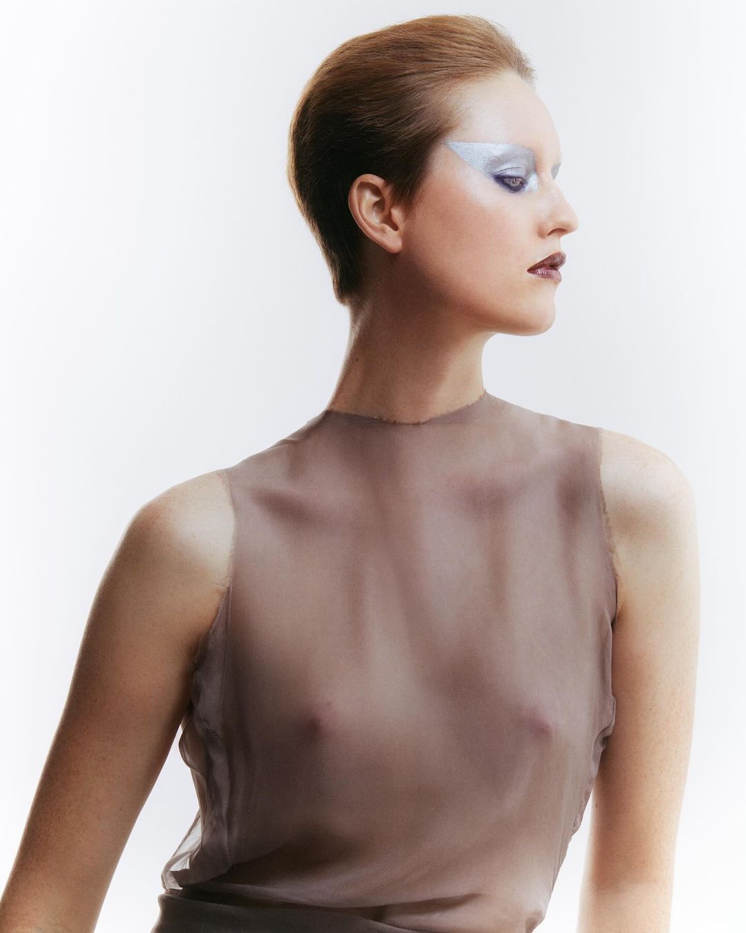 Fashion portrait of an adult woman with short hair wearing a sheer top and bold silver eye makeup against a white background.