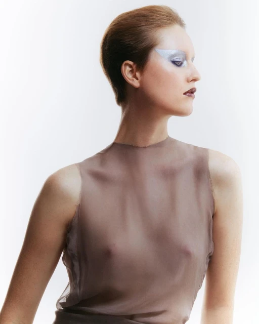 Fashion portrait of an adult woman with short hair wearing a sheer top and bold silver eye makeup against a white background.