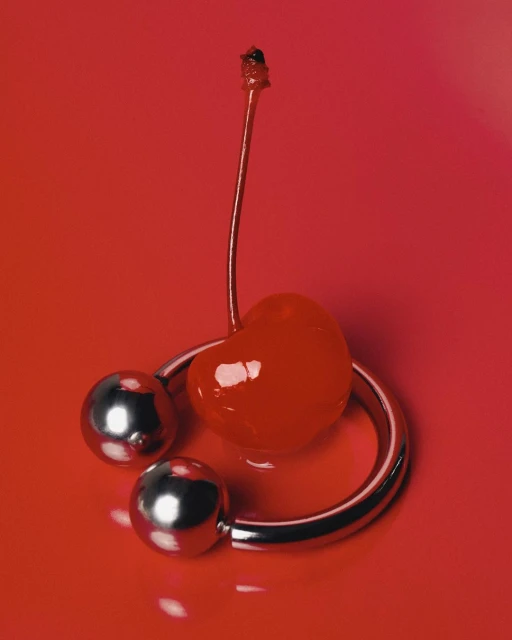 Close-up studio shot of a red cherry next to a metallic, open ring on a saturated red background, showing shiny, minimalist art.
