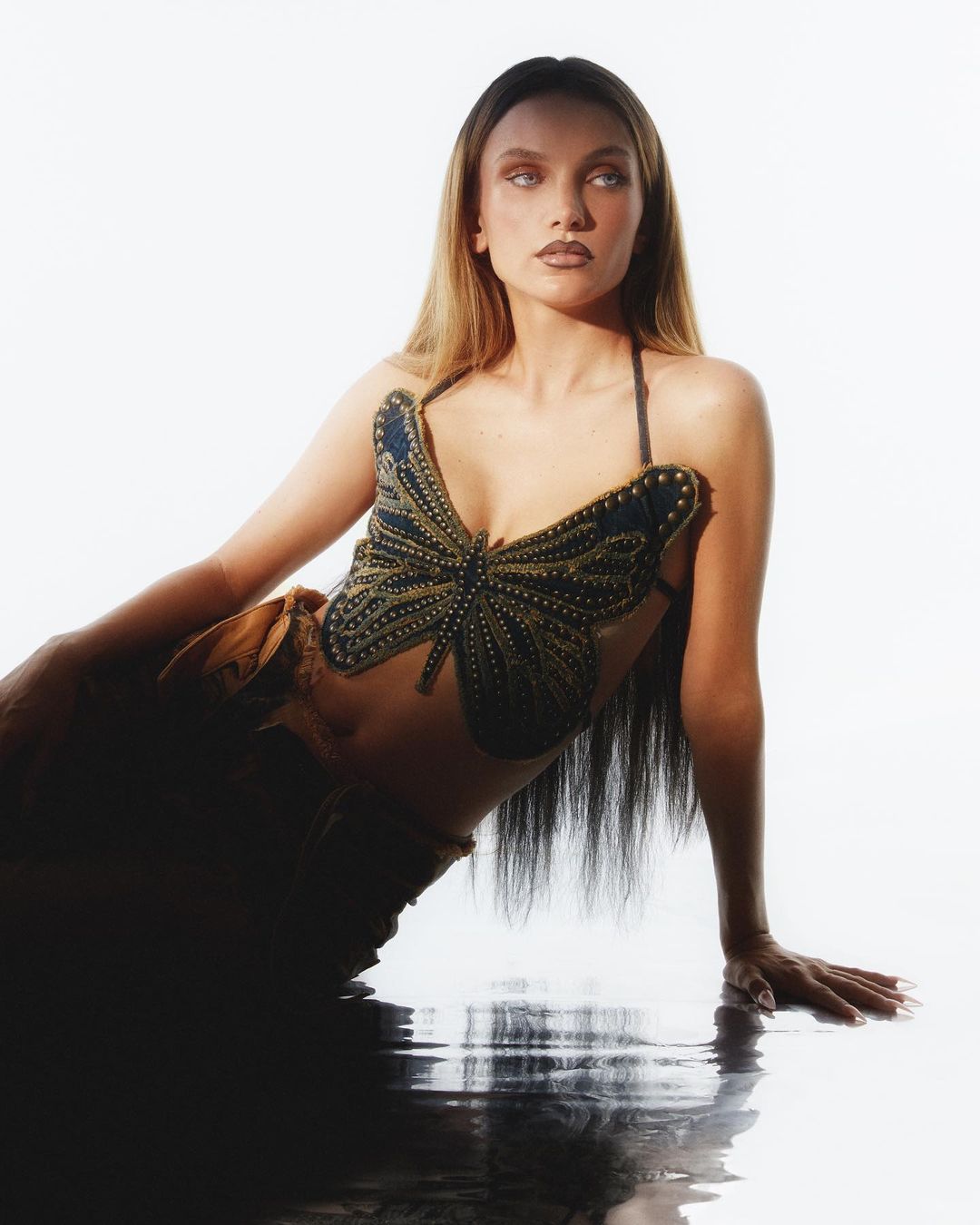 Woman poses in butterfly top and draped skirt against a white backdrop with dark reflection, fashion editorial, studio lighting.