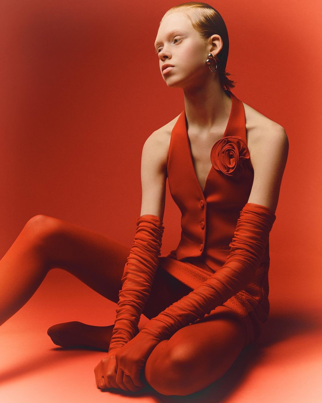 Fashion editorial shot of a fair-skinned young woman in a red vest and leggings, posing against a red backdrop in a studio setting.