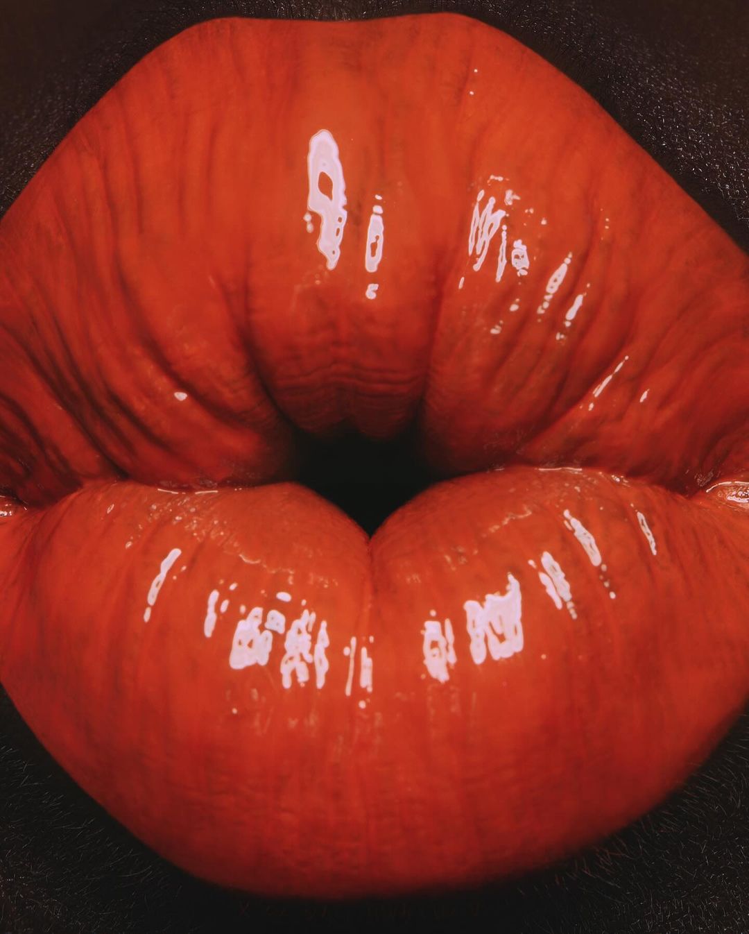 Extreme close-up of full, glossy red lips against a dark skin background, emphasizing detail and color in beauty and fashion context.