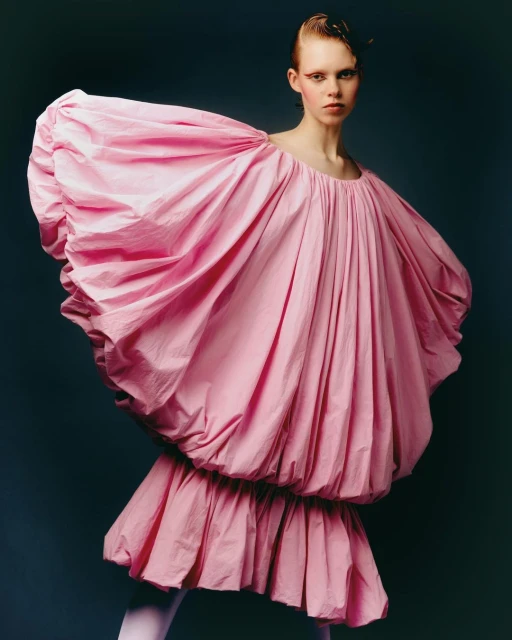 Fashion photography of a young woman posing in a puffy, pink dress and matching stockings, set against a dark background in the studio.