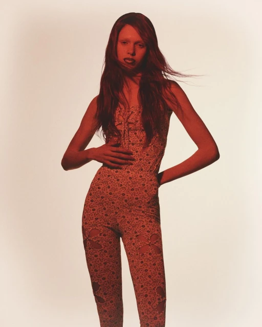 A young adult woman poses in a patterned jumpsuit with her hands on her body. The photo is shot in a studio and has a red color grading.