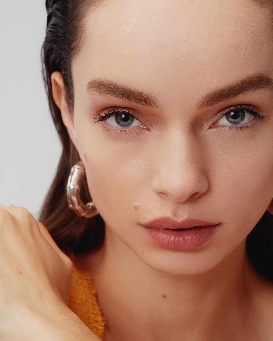 Close-up studio portrait of an adult woman with silver hoop earring, neutral expression, wet hair look, and natural makeup against a white backdrop.