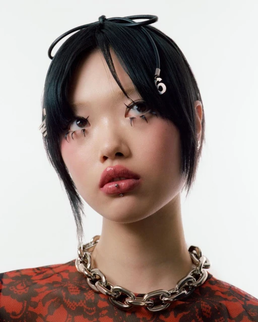 Portrait of a stylish Asian woman with creative makeup, bold jewelry, and an edgy hairstyle against a bright white backdrop.