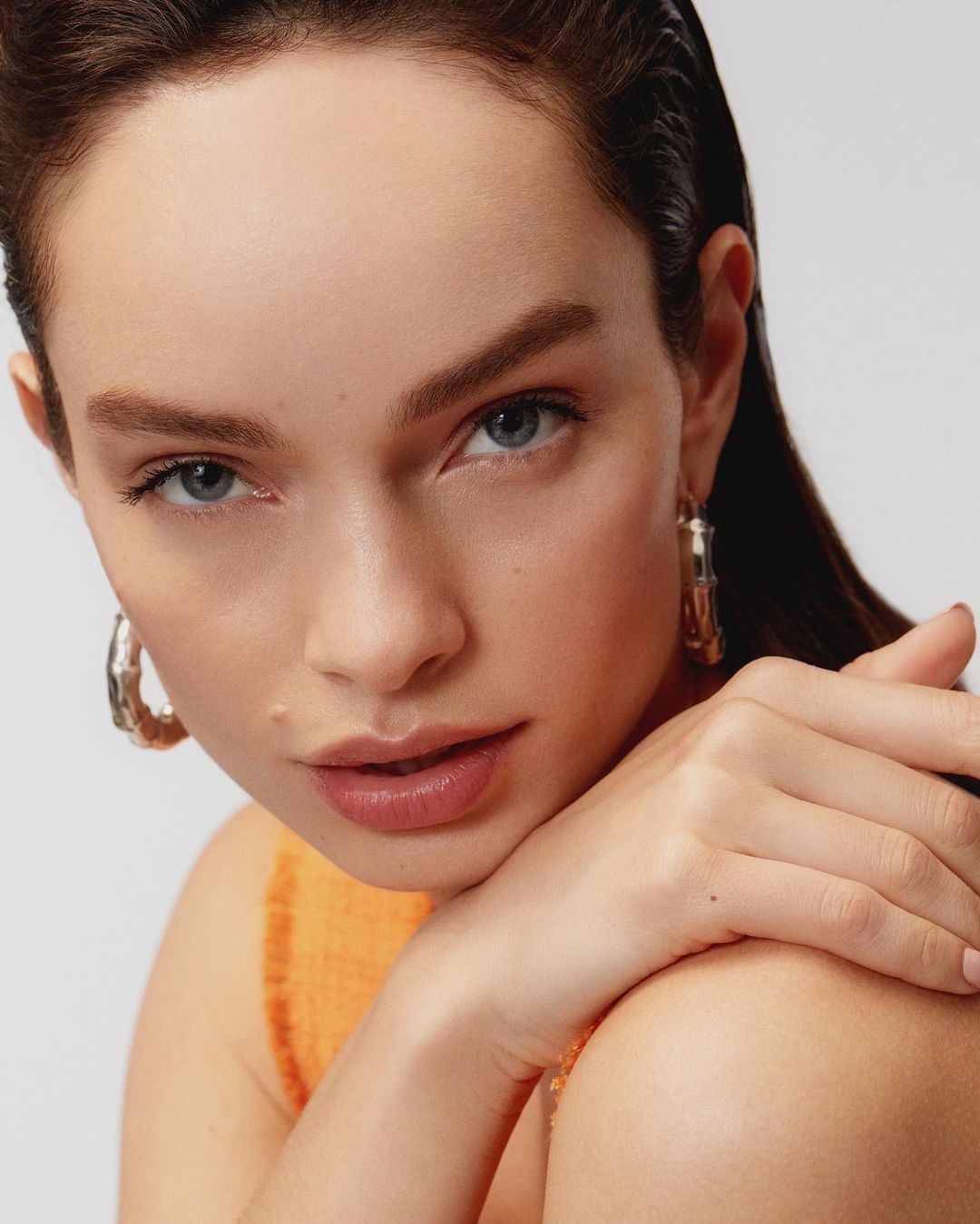 Close-up studio portrait of a woman with hands folded near her face. She has fair skin, blue eyes, and is wearing hoop earrings.