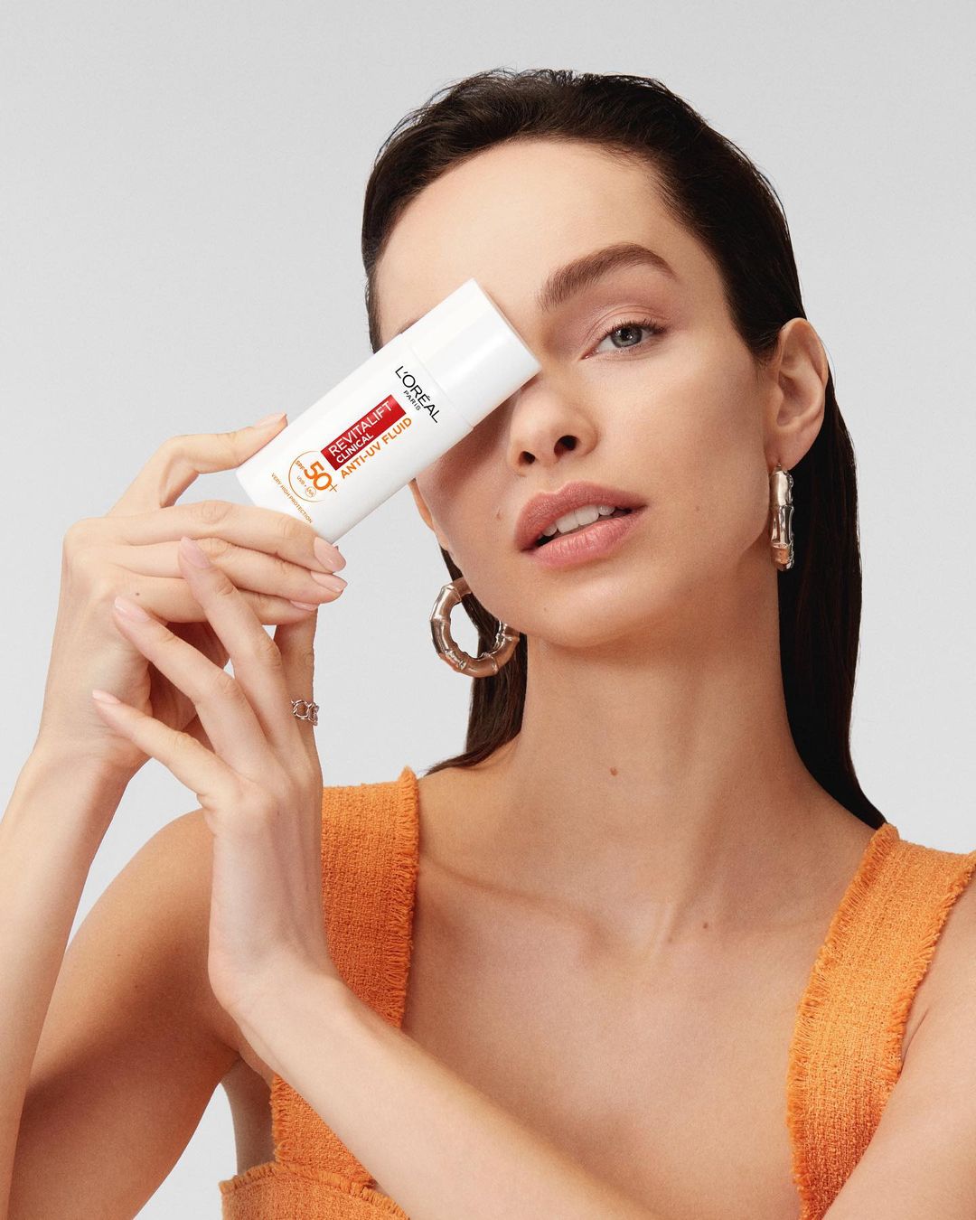 Brunette woman in orange top holding a white tube of L'Oreal Revitalift sunscreen in a studio shot with a neutral background.