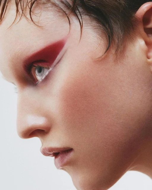 Profile of a woman with bold red graphic eye makeup and slicked-back hair against a clean white background in a beauty shot.