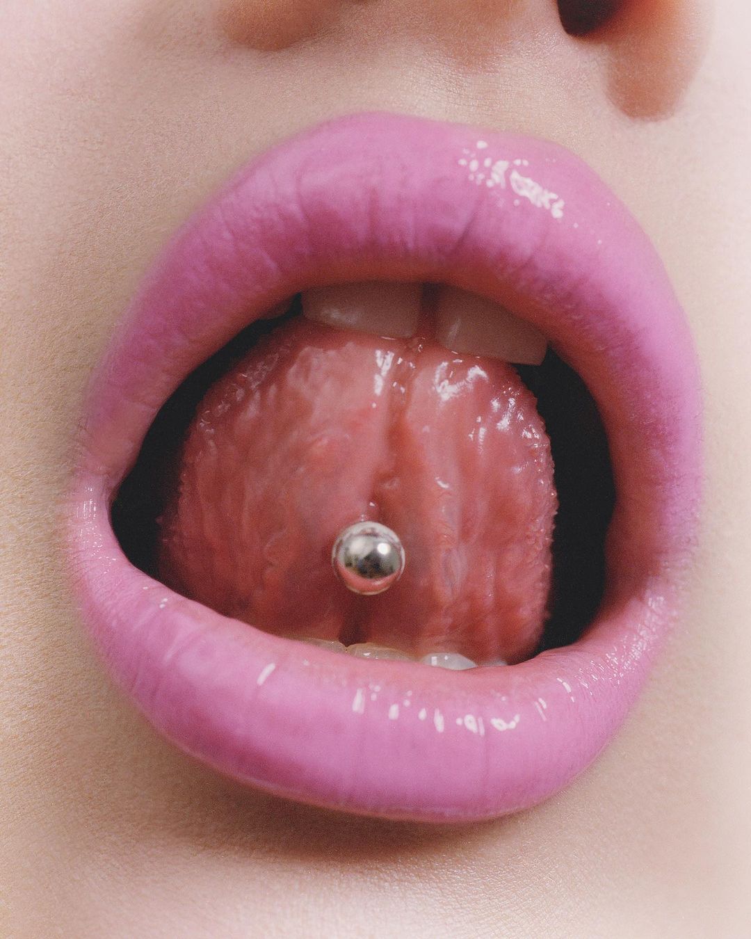 Close-up studio shot of a person's open mouth with pink lipstick, displaying a tongue piercing with a metal ball.