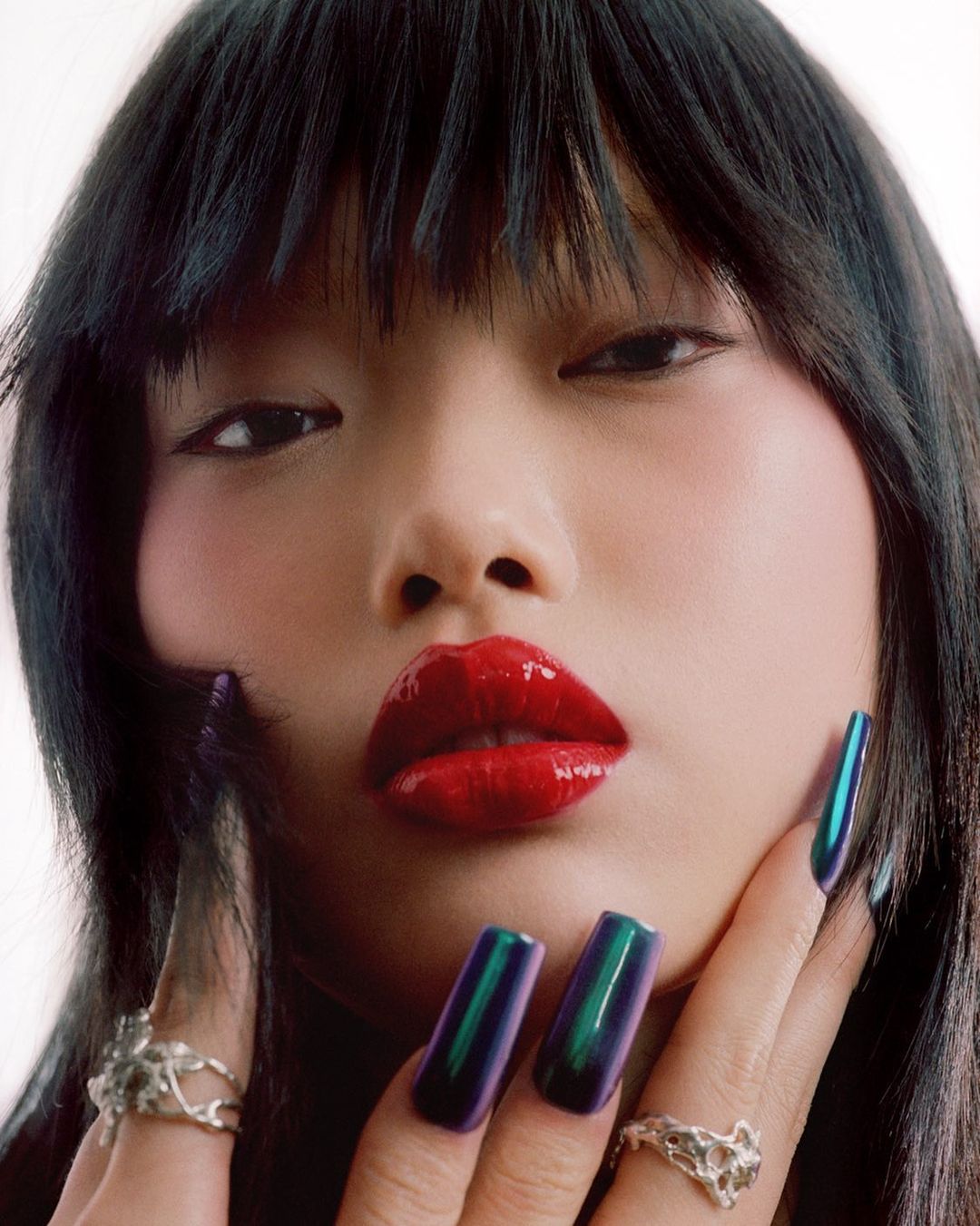 Close-up portrait of a young Asian woman with glossy red lips, iridescent long nails, and silver rings against a bright white background.
