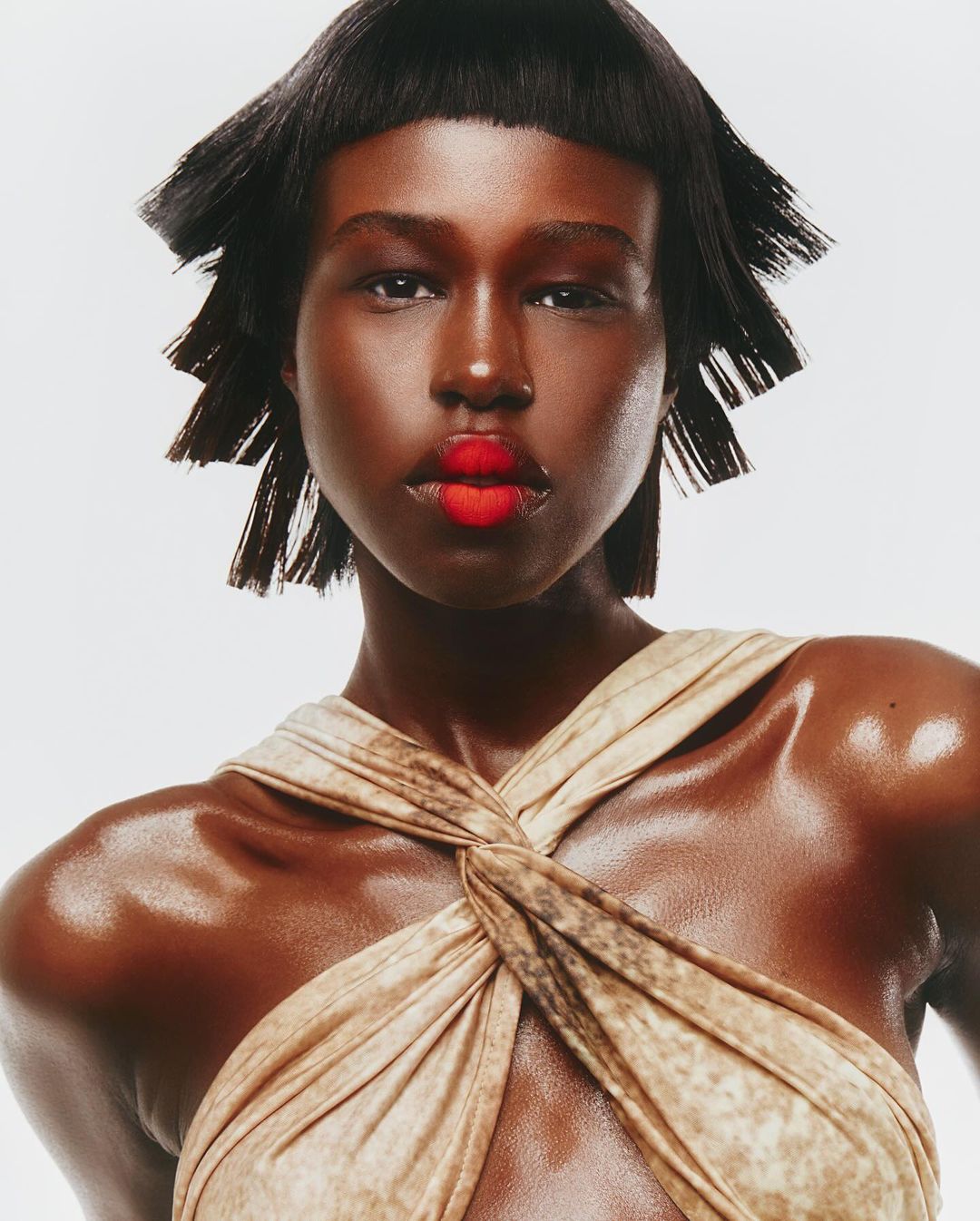 Close-up portrait of a Black female fashion model with short black hair, red lipstick, and a draped tan-colored dress in a studio setting.