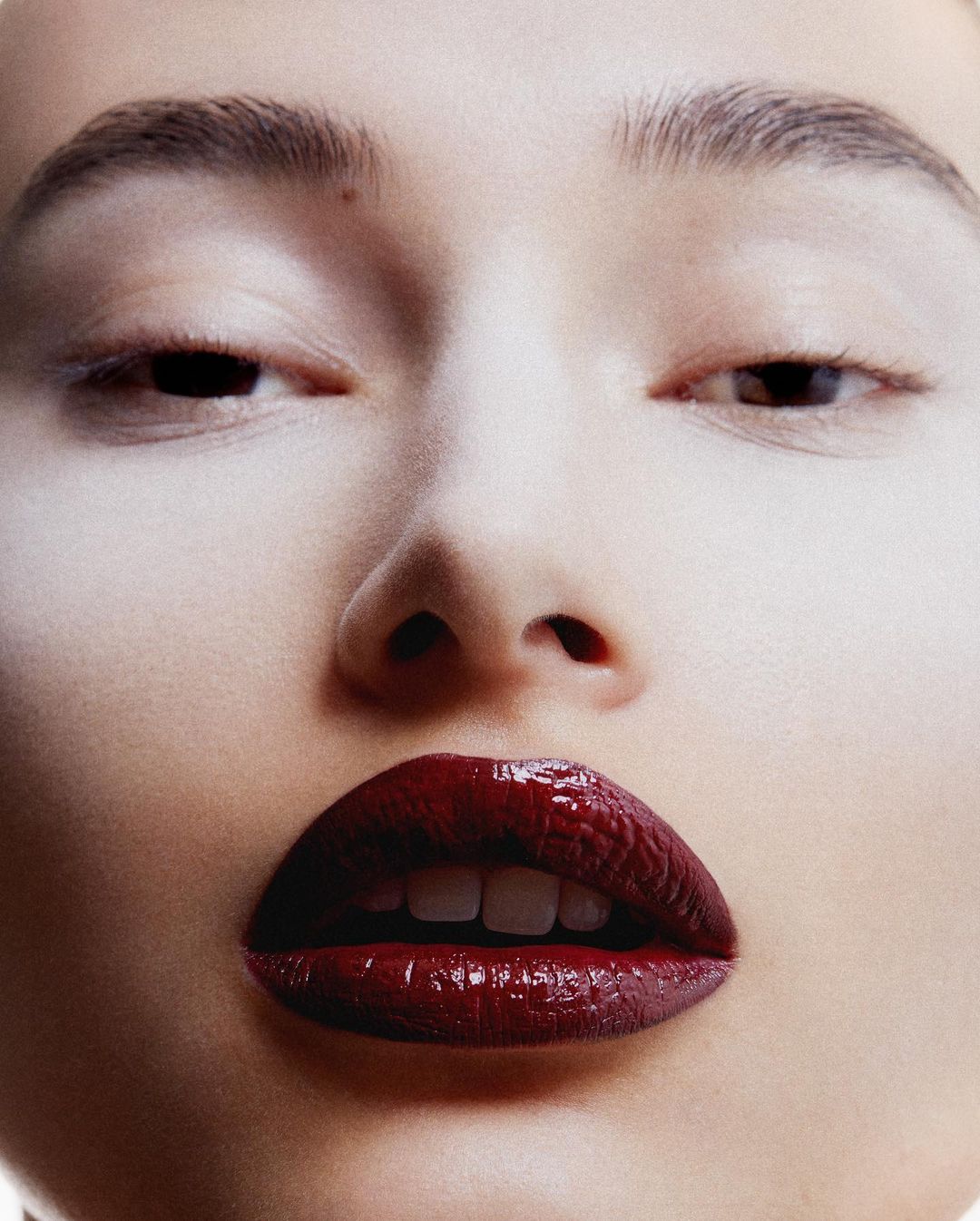 Close-up portrait of a young woman with pale face makeup and dark red glossy lipstick in a beauty and cosmetics context.