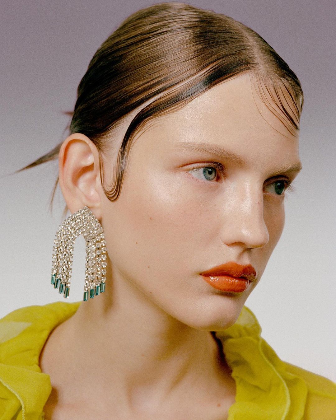 Close-up of a fair-skinned woman with slicked-back brunette hair, orange lipstick, and statement rhinestone earrings, against a neutral backdrop.