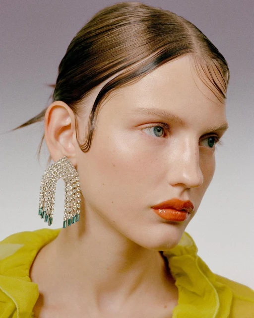 Close-up of a fair-skinned woman with slicked-back brunette hair, orange lipstick, and statement rhinestone earrings, against a neutral backdrop.