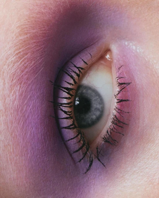 Close-up shot of a single eye with long eyelashes, pink and purple lighting, and smooth skin texture in a beauty-focused composition.
