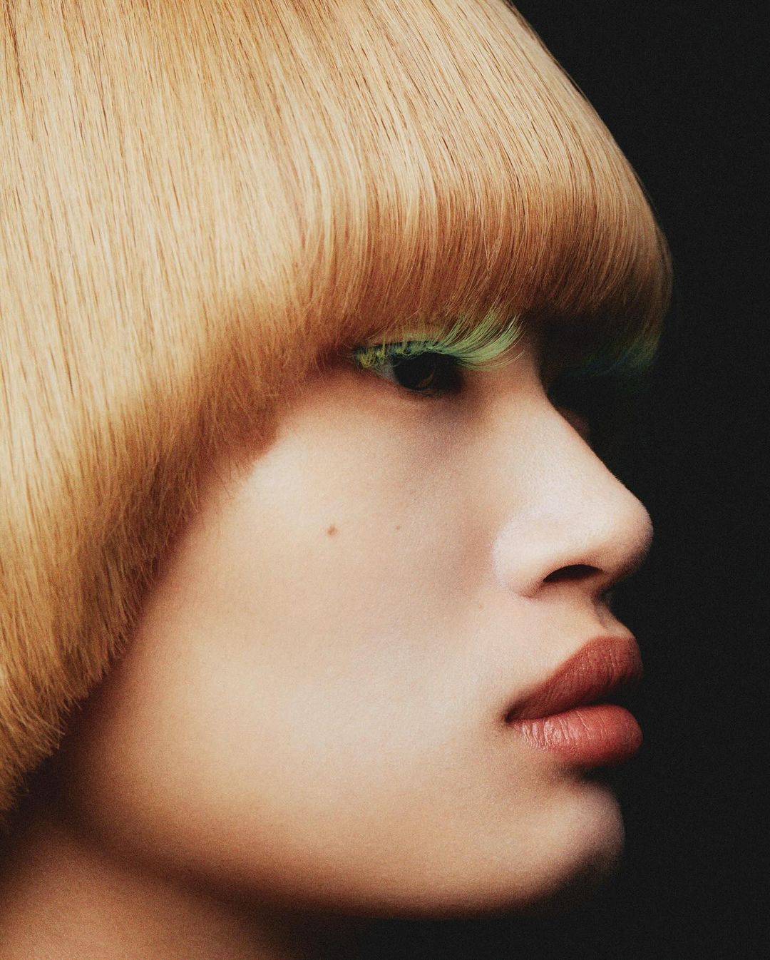 Close-up of a person with fair skin, blunt blonde bob, and green eyelashes against a dark background. Focus on makeup and hair.
