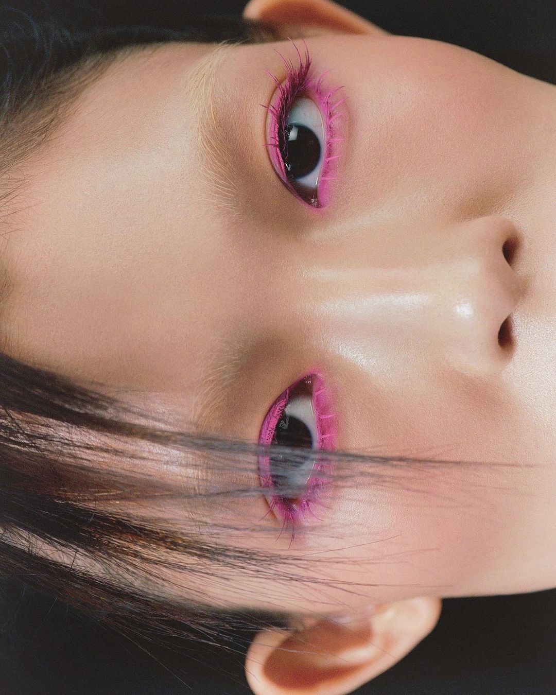Close-up portrait of a person with pink makeup around the eyes and dark hair against a dark backdrop, showcasing beauty and style.