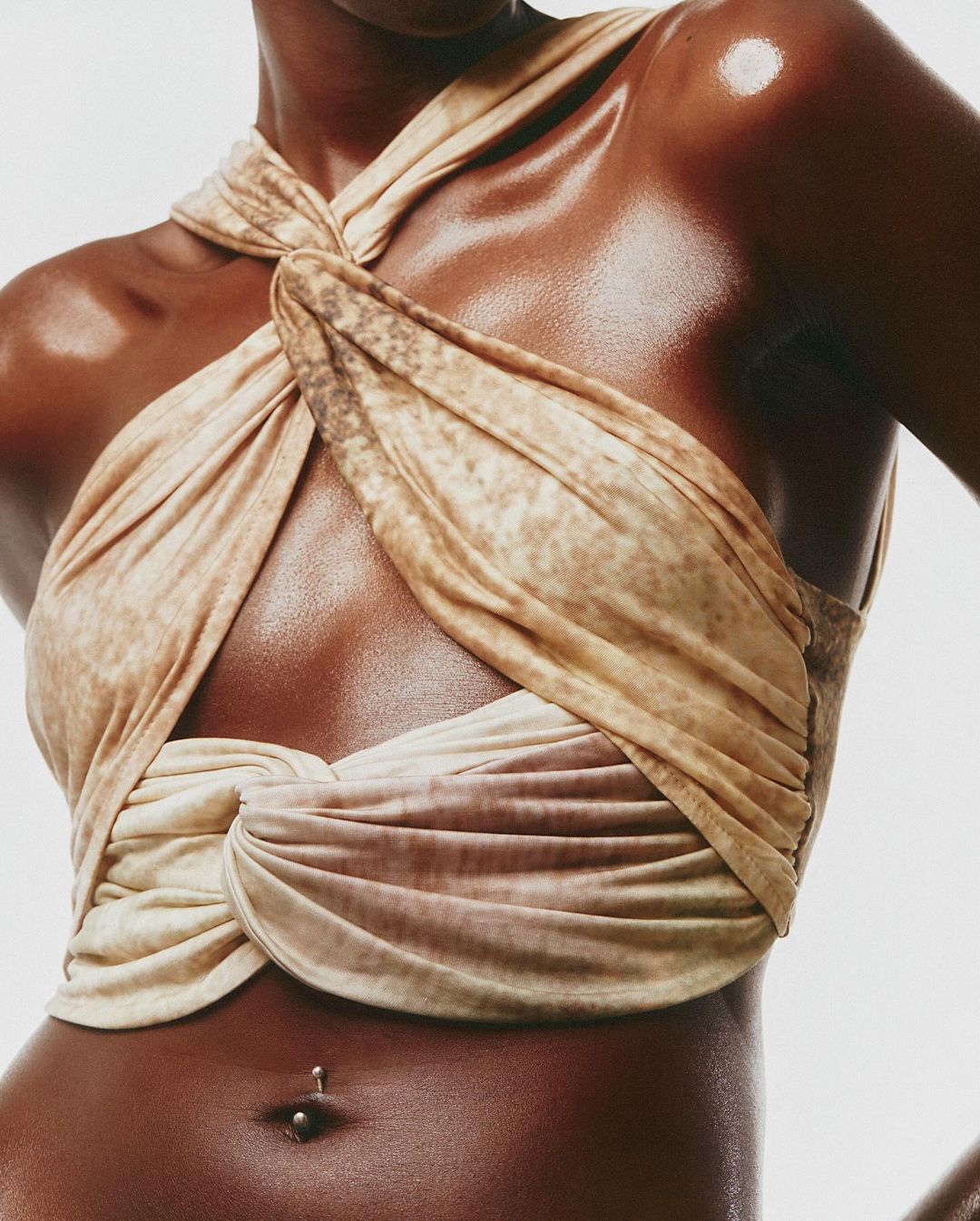 Close-up studio shot of a tanned adult female with a belly ring, wearing a beige twisted fabric top against a bright background.