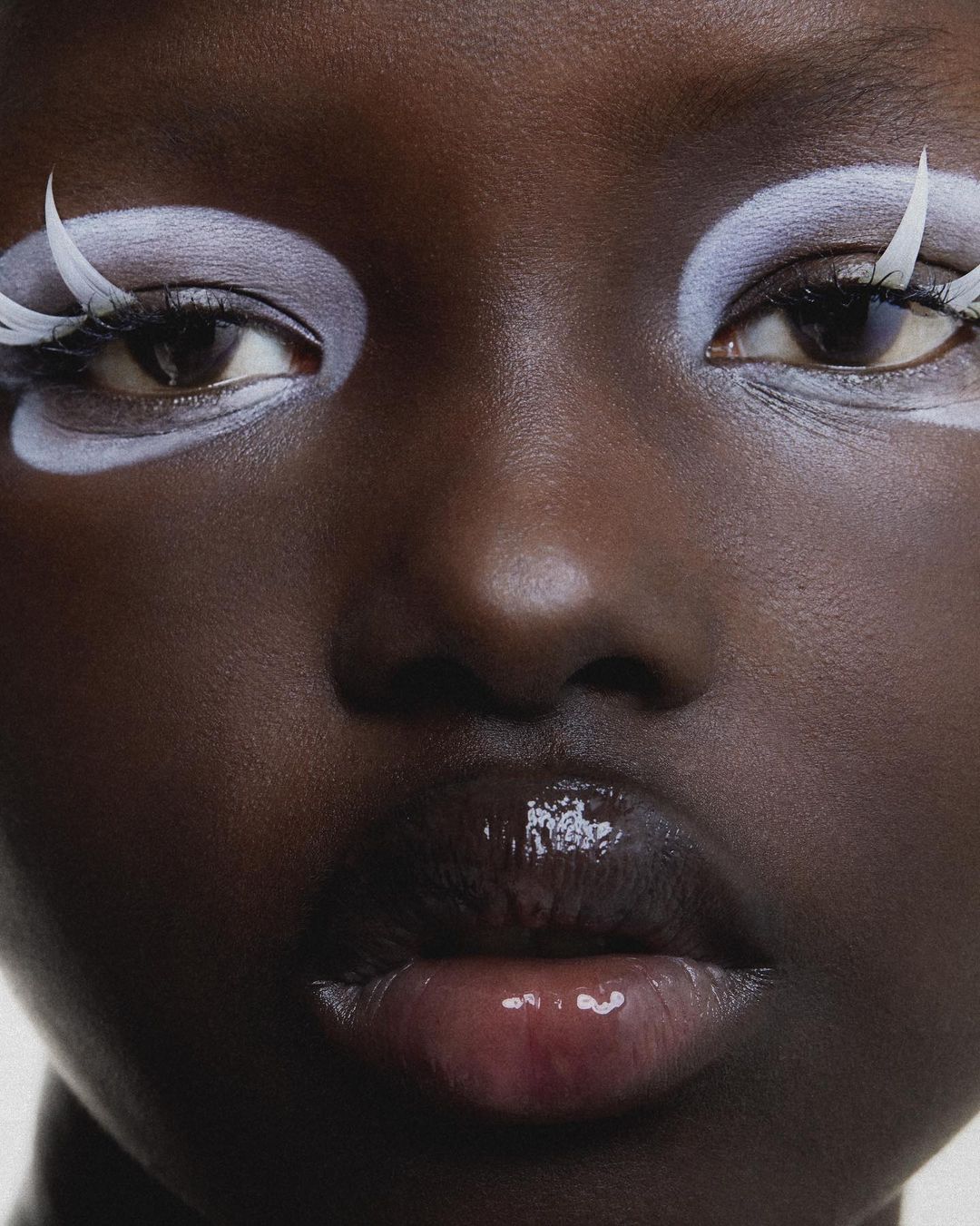 Close-up portrait of a woman with dark skin and glossy lips. She has dramatic white makeup on her eyes and eyebrows, along with white lashes.