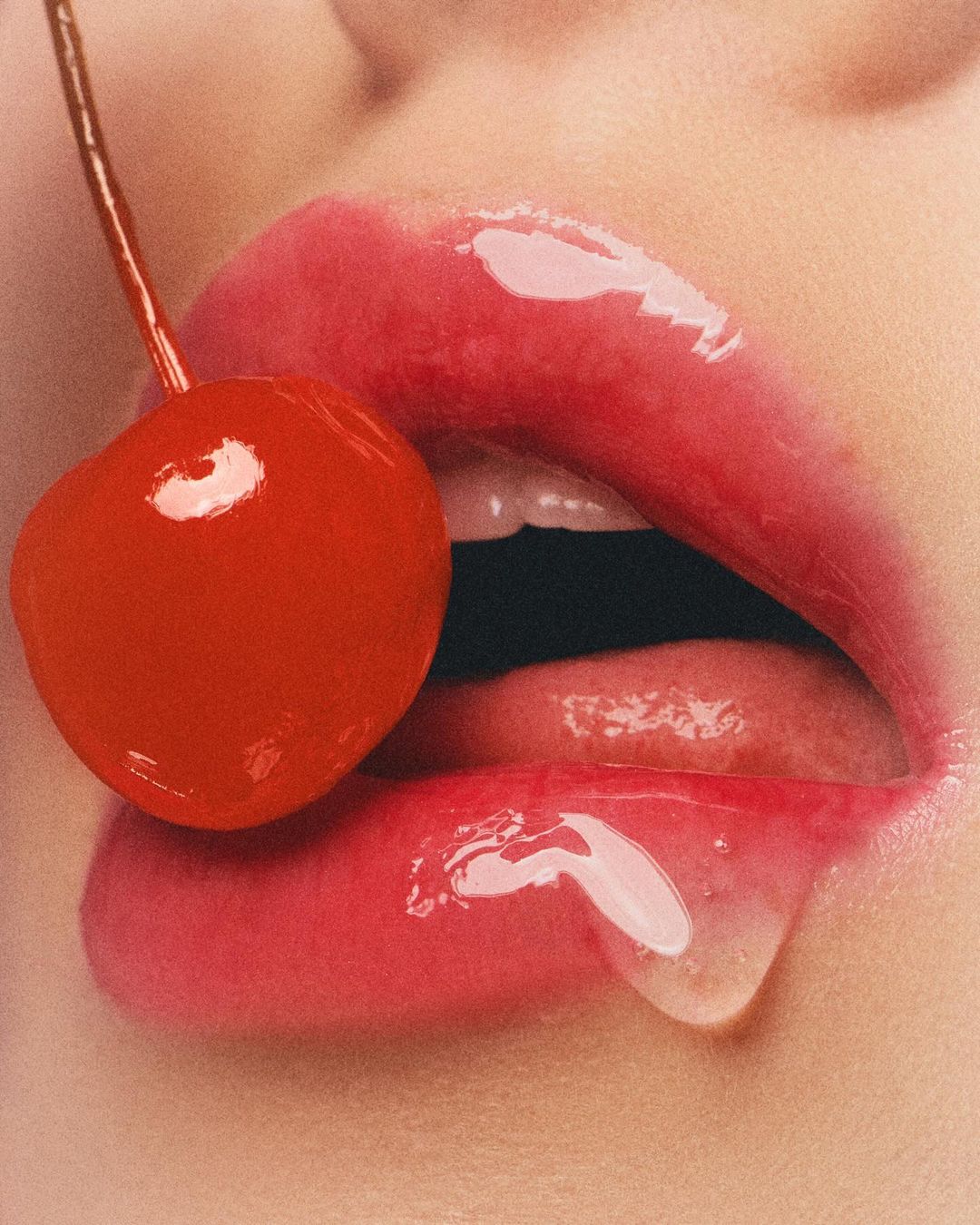 Close-up of a woman's glossy lips with a bright red cherry held to them. The cherry and lips dominate the close-up composition.