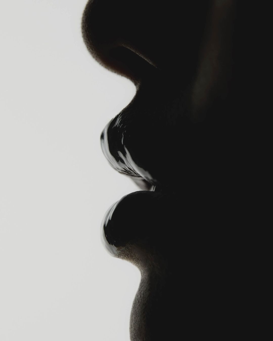 Close-up profile of a human face, with dark glossy makeup or paint covering the lips and skin, set against a plain white background.