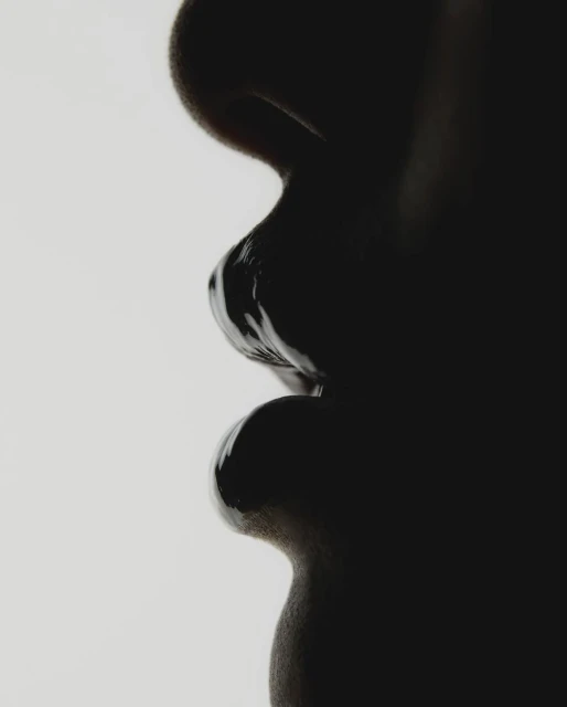 Close-up profile of a human face, with dark glossy makeup or paint covering the lips and skin, set against a plain white background.
