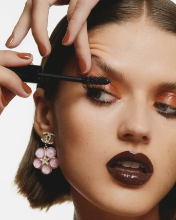 Close-up beauty shot of a woman having mascara applied to her eyelashes by a manicured hand, wearing statement earrings.