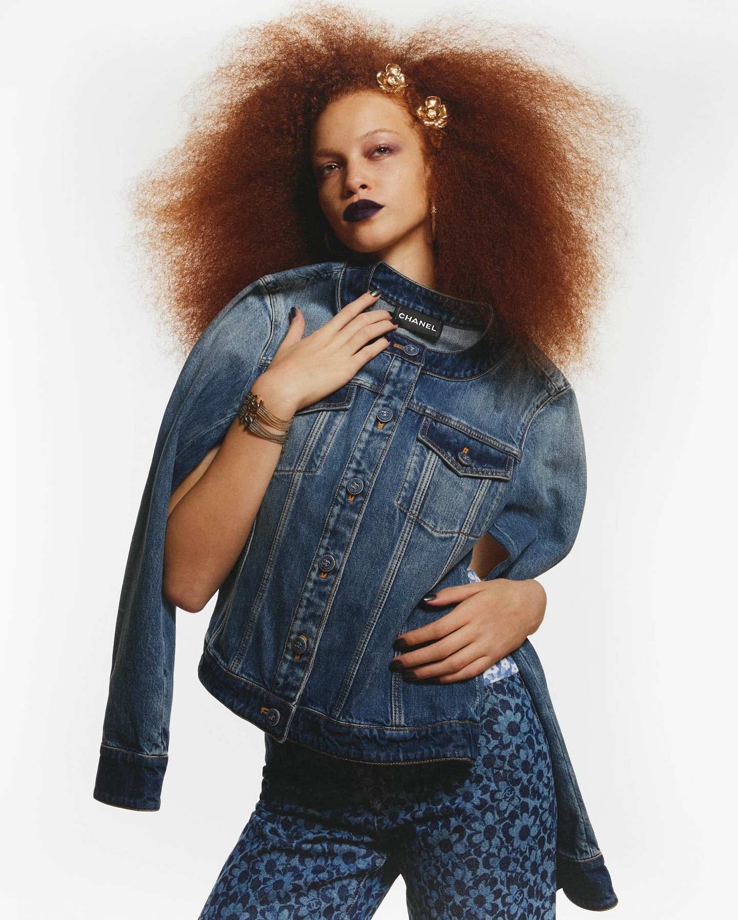 A stylish red-haired woman poses indoors wearing a denim jacket and floral pants, complemented by dark lipstick and flower hair accessories.
