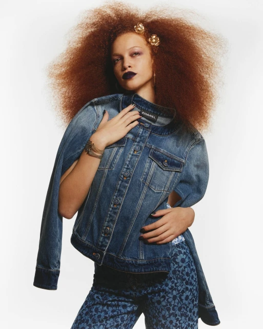 A stylish red-haired woman poses indoors wearing a denim jacket and floral pants, complemented by dark lipstick and flower hair accessories.