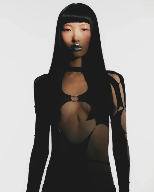 Asian woman poses in a stylish black cutout dress, silver makeup, and long black hair against a white background in a fashion shot.