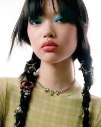 Close-up portrait of a model with braided dark hair, blue eyeshadow, and silver jewelry captured against a bright white background.