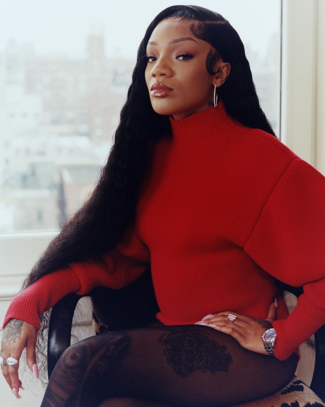 Portrait of a Black woman with long dark hair wearing a red turtleneck sweater, sitting on a chair in front of a window with a city view.