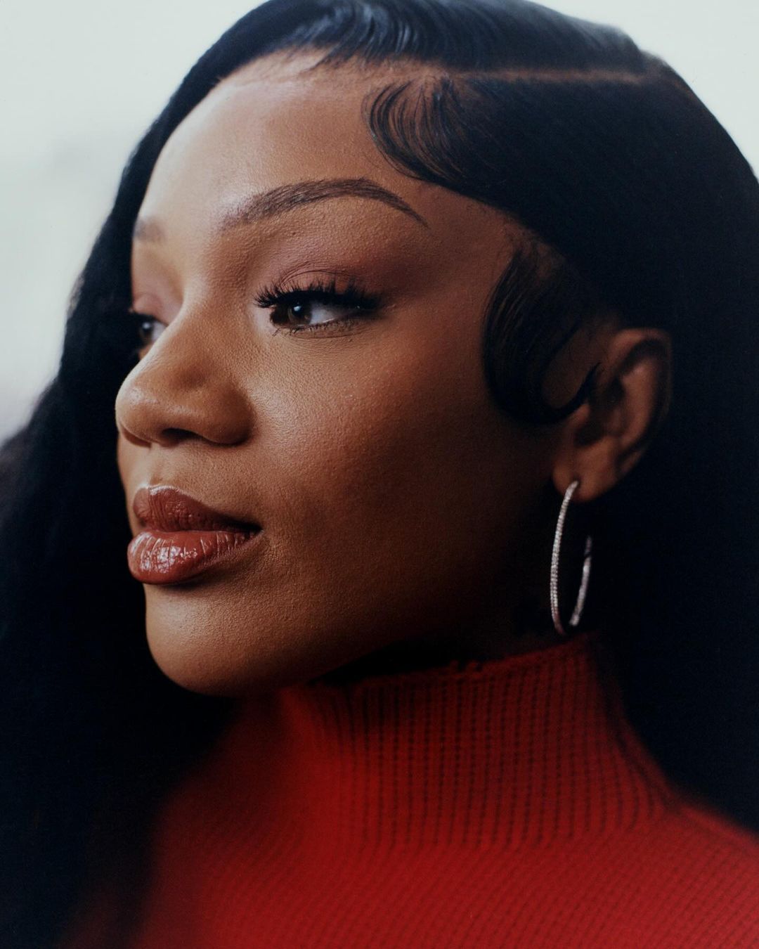 Close-up portrait of a woman with dark hair and red turtleneck looking off to the side with makeup and hoop earrings against soft lighting.