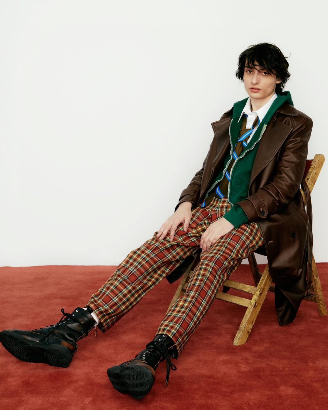 A young adult poses on a wooden chair in a brown leather coat, green sweater, tie, and plaid pants against a white backdrop.