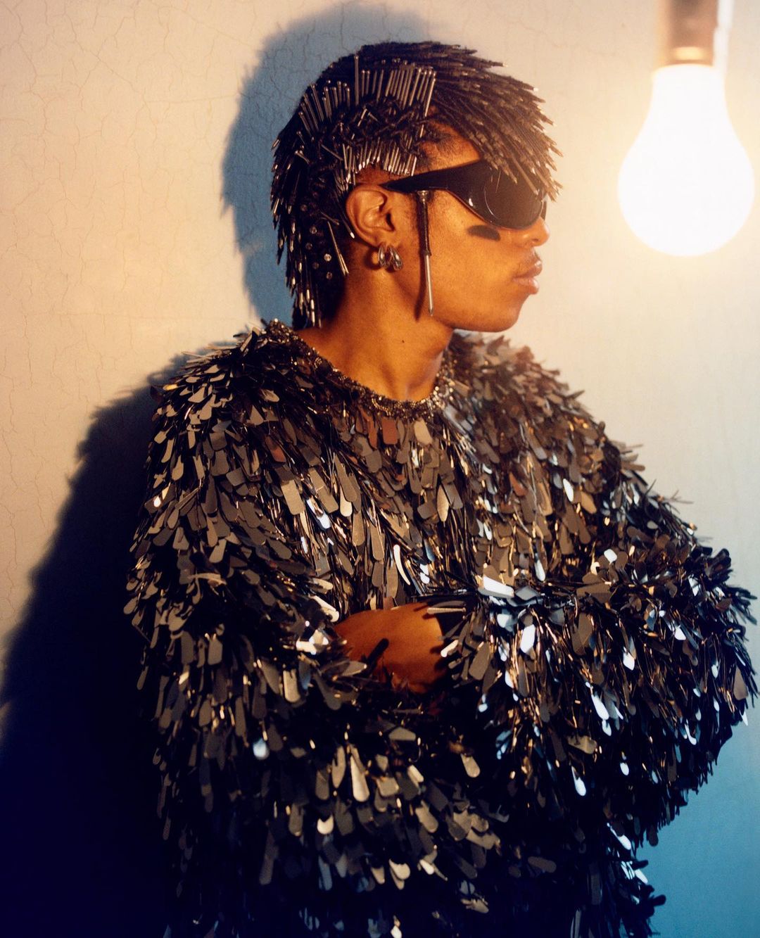 Profile of person wearing sunglasses and a silver sequin top standing indoors with a lightbulb and grey wall in the background.