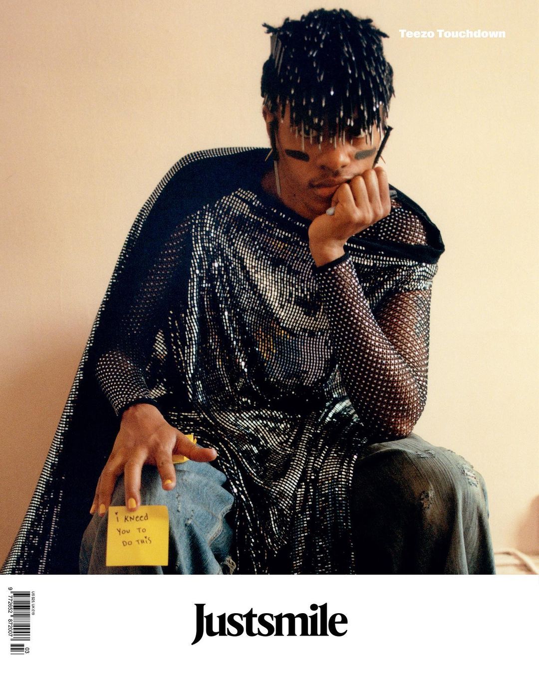 Stylish man in a shimmering outfit sits pensively against a plain wall with handwritten notes on jeans. Editorial fashion portrait.