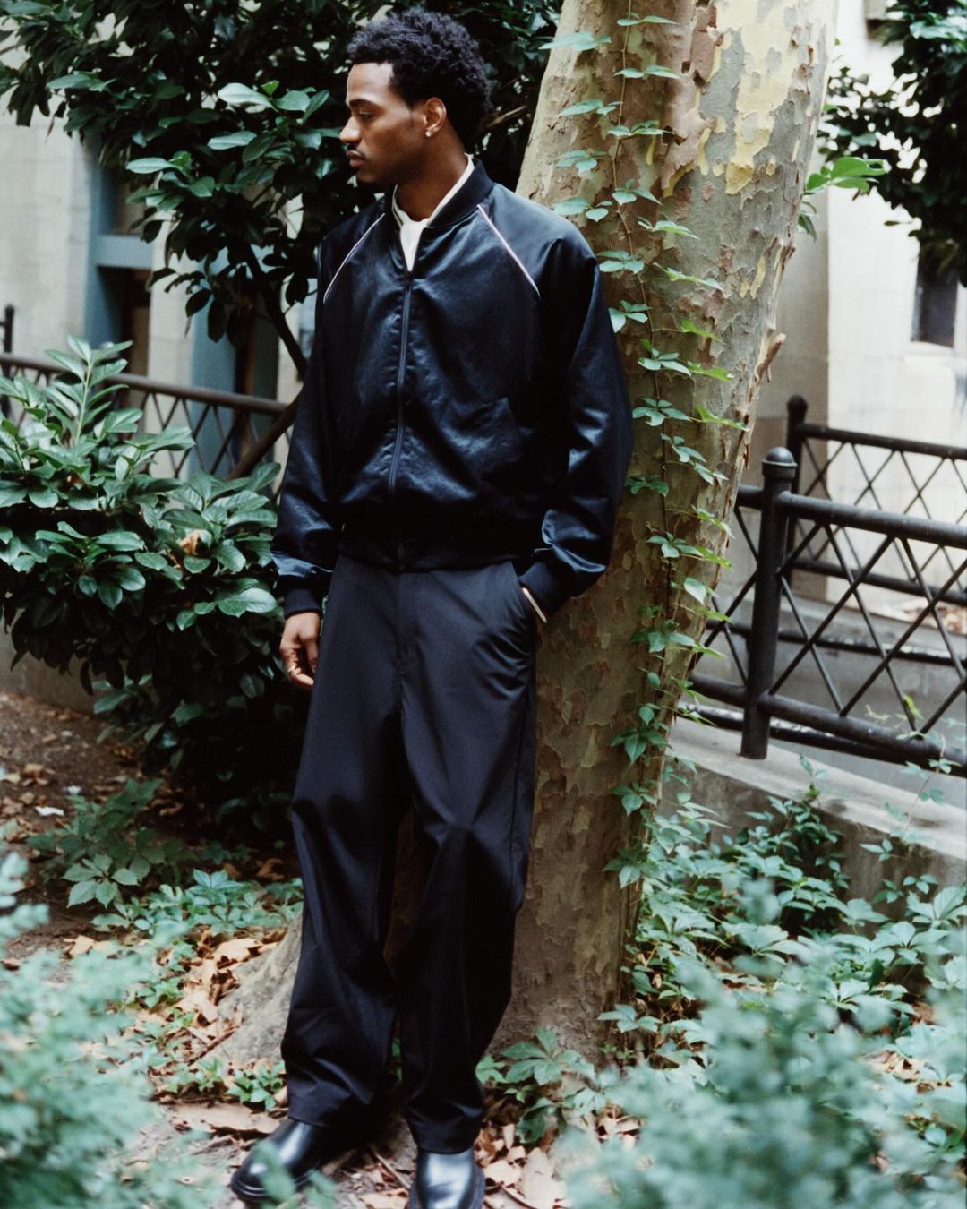 An African-American man stands near a tree in an urban environment wearing a black bomber jacket, black pants, and shiny black shoes.