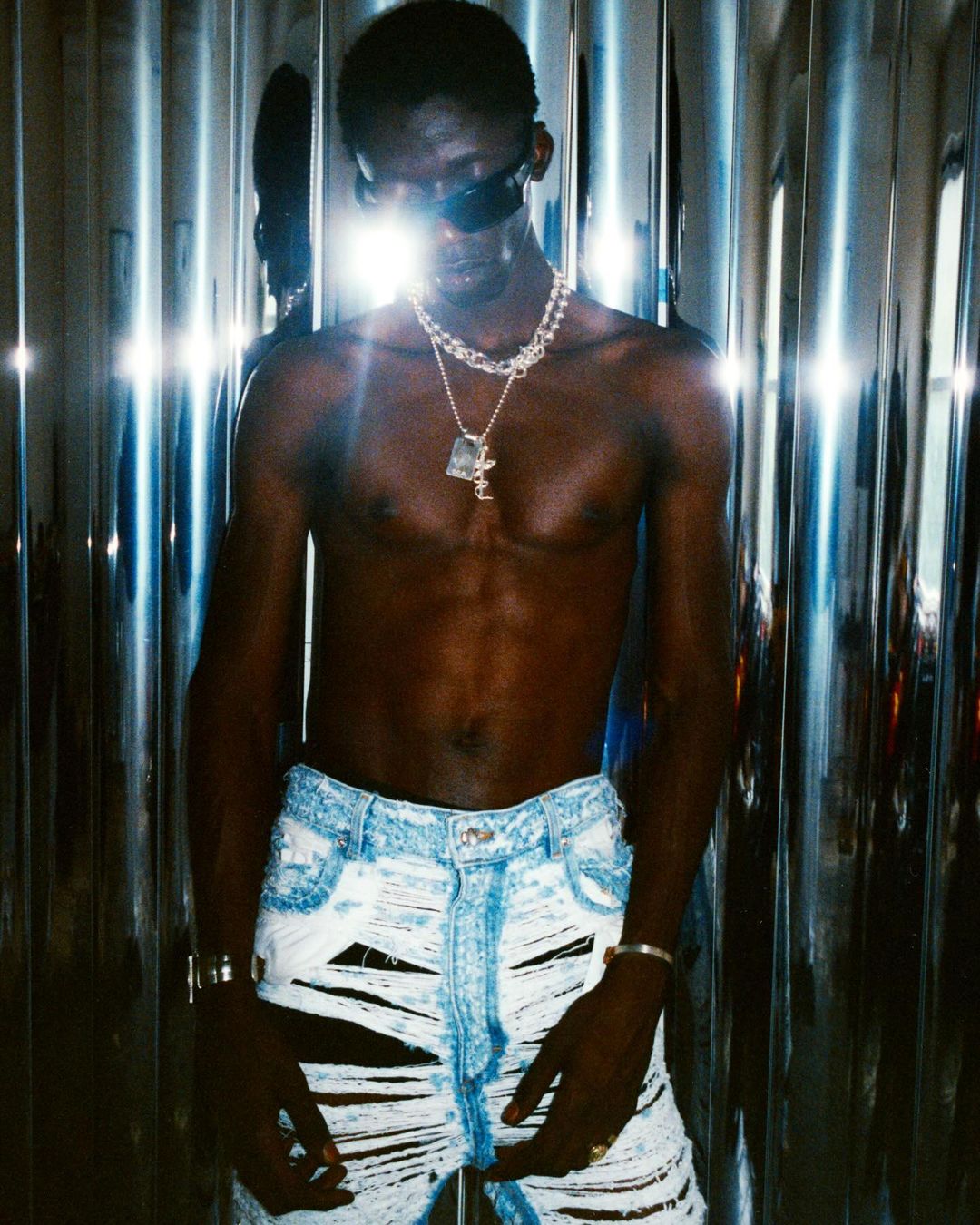 A shirtless African male model poses in ripped jeans and sunglasses against a mirrored wall, highlighted by bright studio lighting.