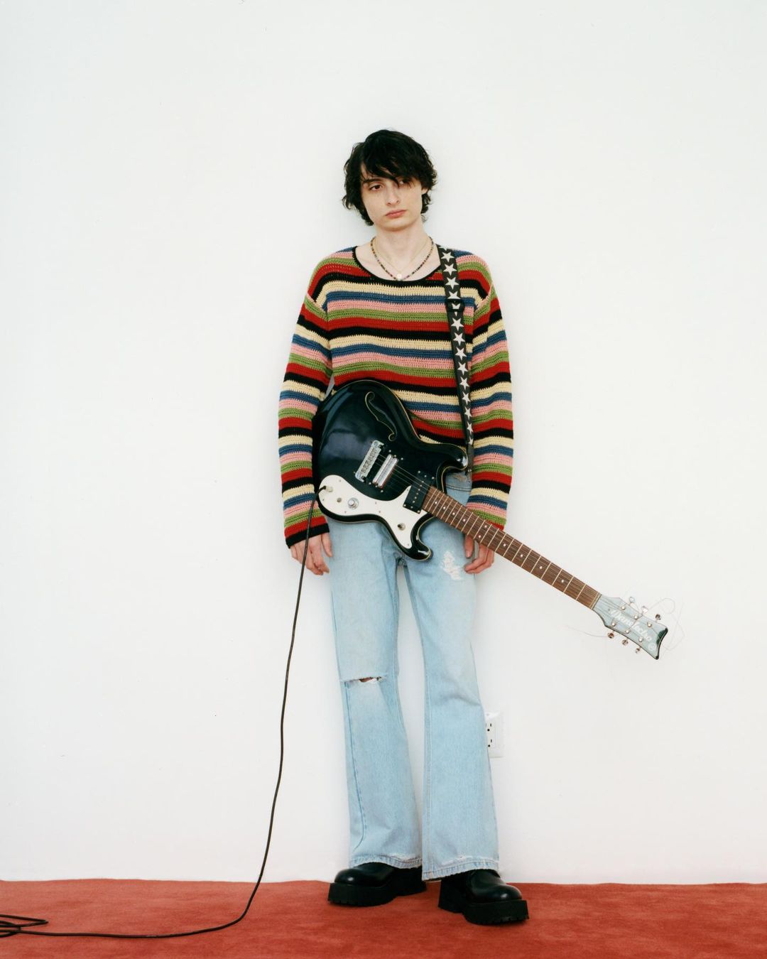 Young adult stands indoors against a white wall, wearing a striped sweater, ripped jeans, black boots, and holding an electric guitar.