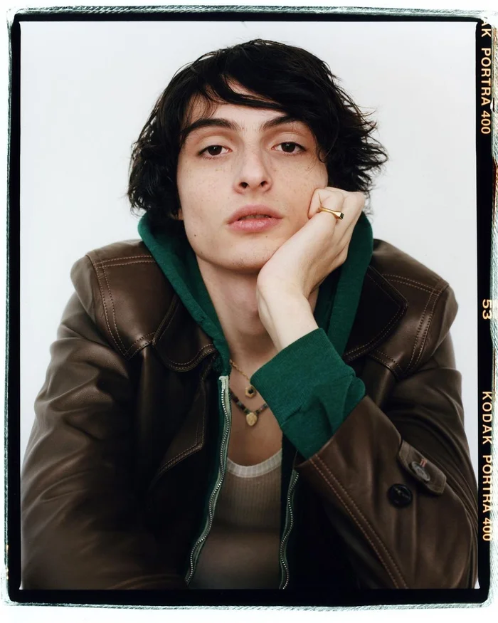 Portrait of young man with dark hair in brown leather jacket and green hoodie resting chin on hand against white background
