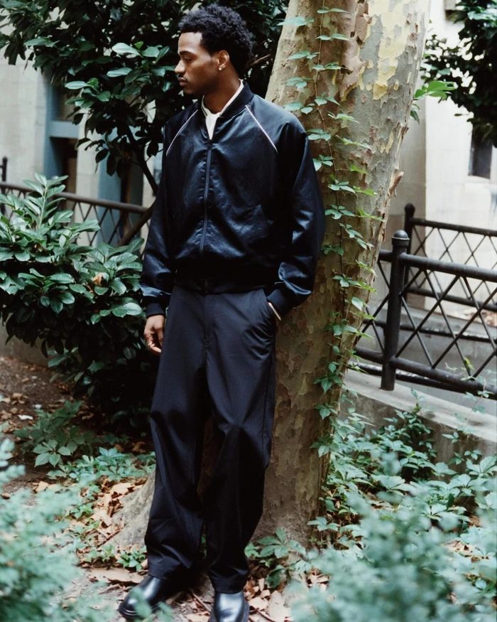 Young Black man wearing a black bomber jacket and dark trousers standing beside a tree in New York by Julius Frazer