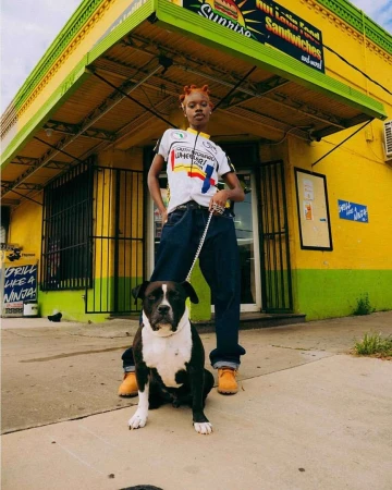 A young adult woman stands with a black and white dog in front of a vibrant yellow and green building on a sunny day.