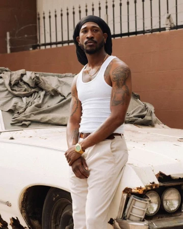 Adult male with dark hair poses in front of a vintage white car, wearing a white tank top, gold watch, and tan pants.