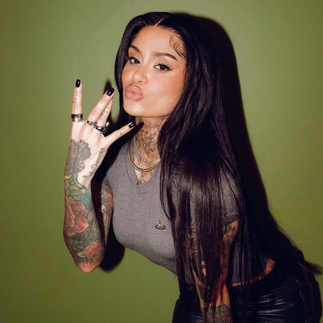 A woman with tattoos poses against a green wall. She is wearing a grey shirt, black pants, and jewelry, and is making a hand gesture.
