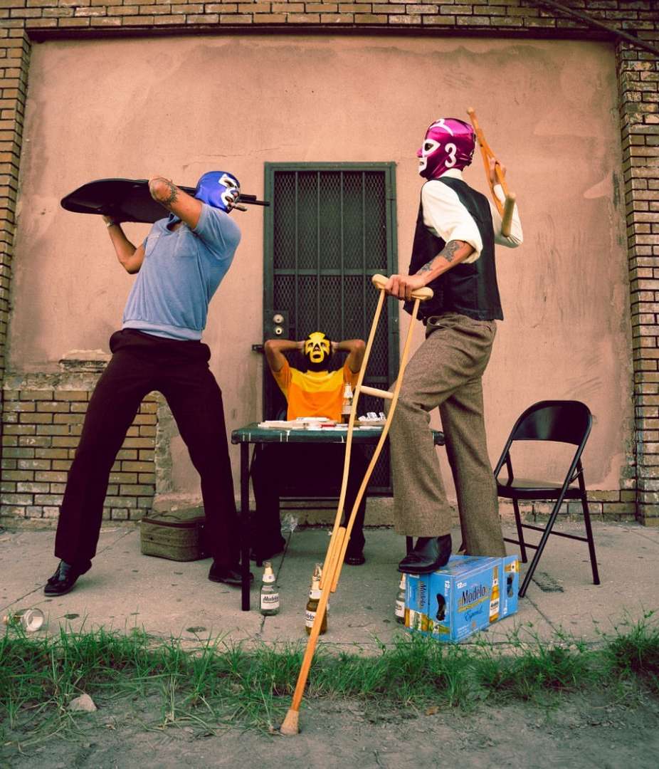 Outdoor photo of three luchadores in masks; one holds a tray, another leans on a crutch by beers, and the third sits at a table.