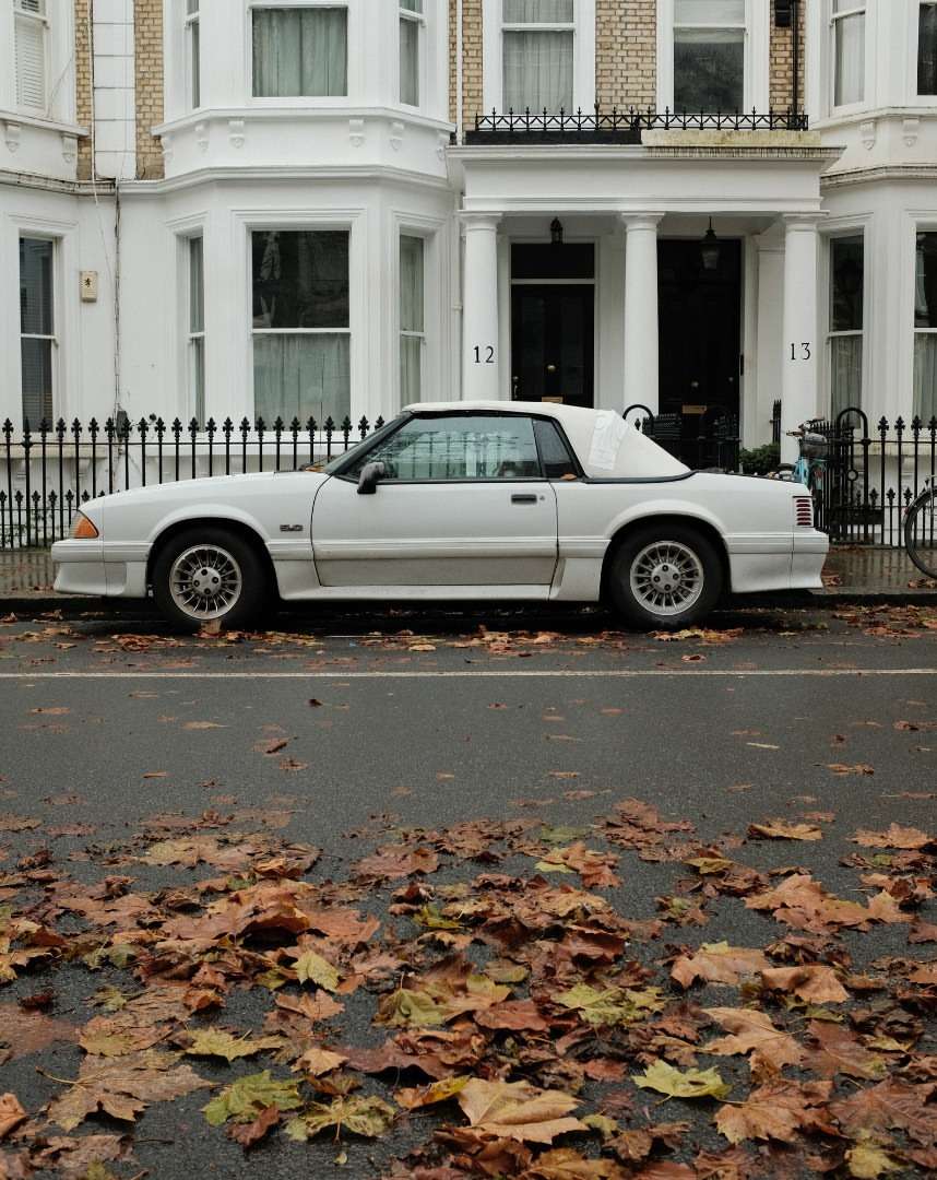 White convertible car parked on a street in front of white townhouses, leaves scattered on the wet pavement, overcast day.