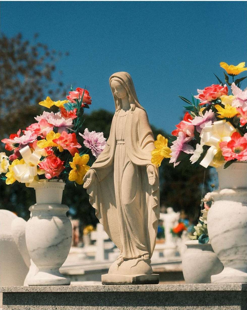 Outdoor shot of a stone Virgin Mary statue, draped with colorful flowers, set within a graveyard under a bright blue sky, close-up view.