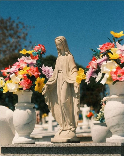 Outdoor shot of a stone Virgin Mary statue, draped with colorful flowers, set within a graveyard under a bright blue sky, close-up view.