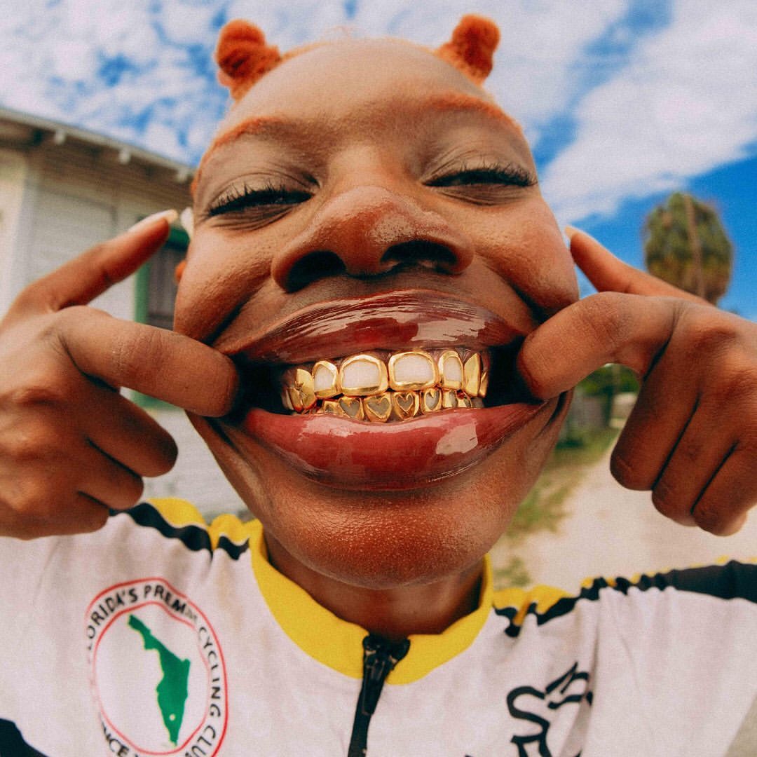Close-up of a Black woman with orange hair pulling her lips back to show off her gold grill with heart-shaped designs outdoors.